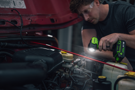 A mechanic wearing safety glasses works under a red car hood, using a green and black cordless flashlight to inspect the engine components.