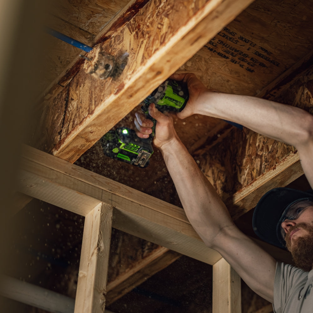 A man using a green and black cordless drill in a wooden attic or construction setting.