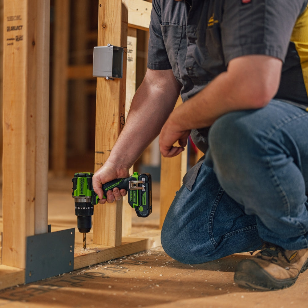 A construction worker kneels on a wooden floor, using a green and black cordless drill to fasten a metal bracket to a wooden framing post in an unfinished building.