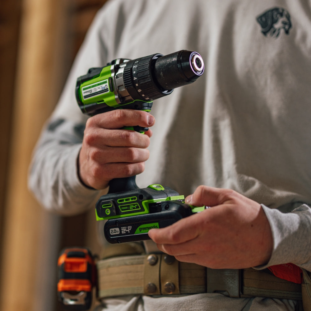A person in a light gray long-sleeve shirt holds a green and black cordless drill in both hands, with a work belt and orange tool visible at the waist. The background is a softly blurred workshop setting.