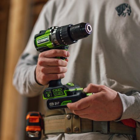 A person in a light gray long-sleeve shirt holds a green and black cordless drill in both hands, with a work belt and orange tool visible at the waist. The background is a softly blurred workshop setting.